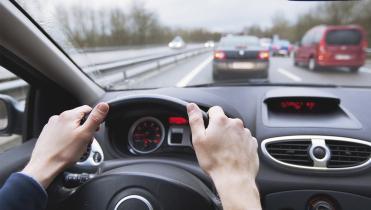 Two hands on the steering wheel of a car with a view of the road ahead.